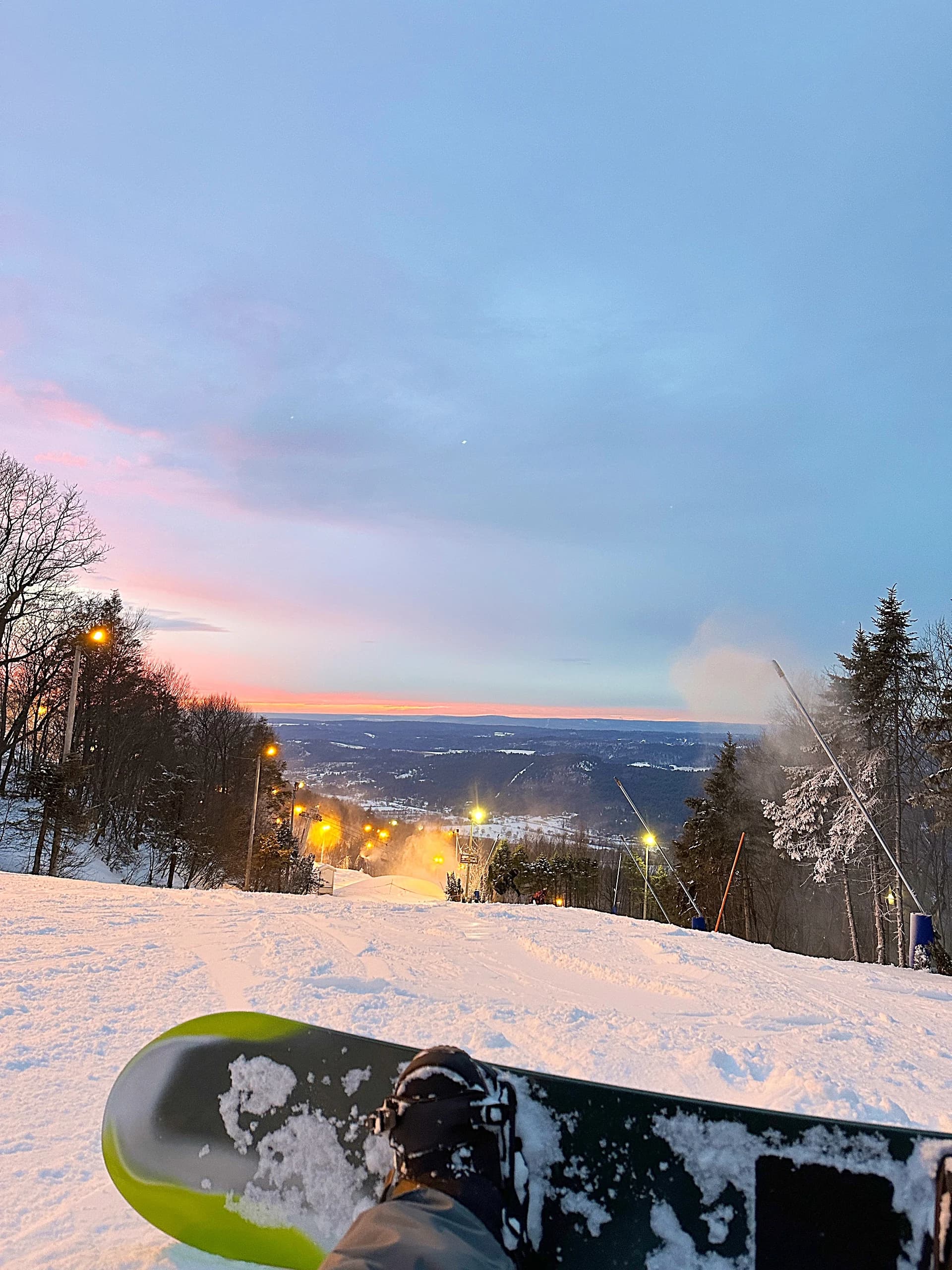 The nose of a snowboard in the foreground, looking down a floodlit slope toward a pink dusk sky.